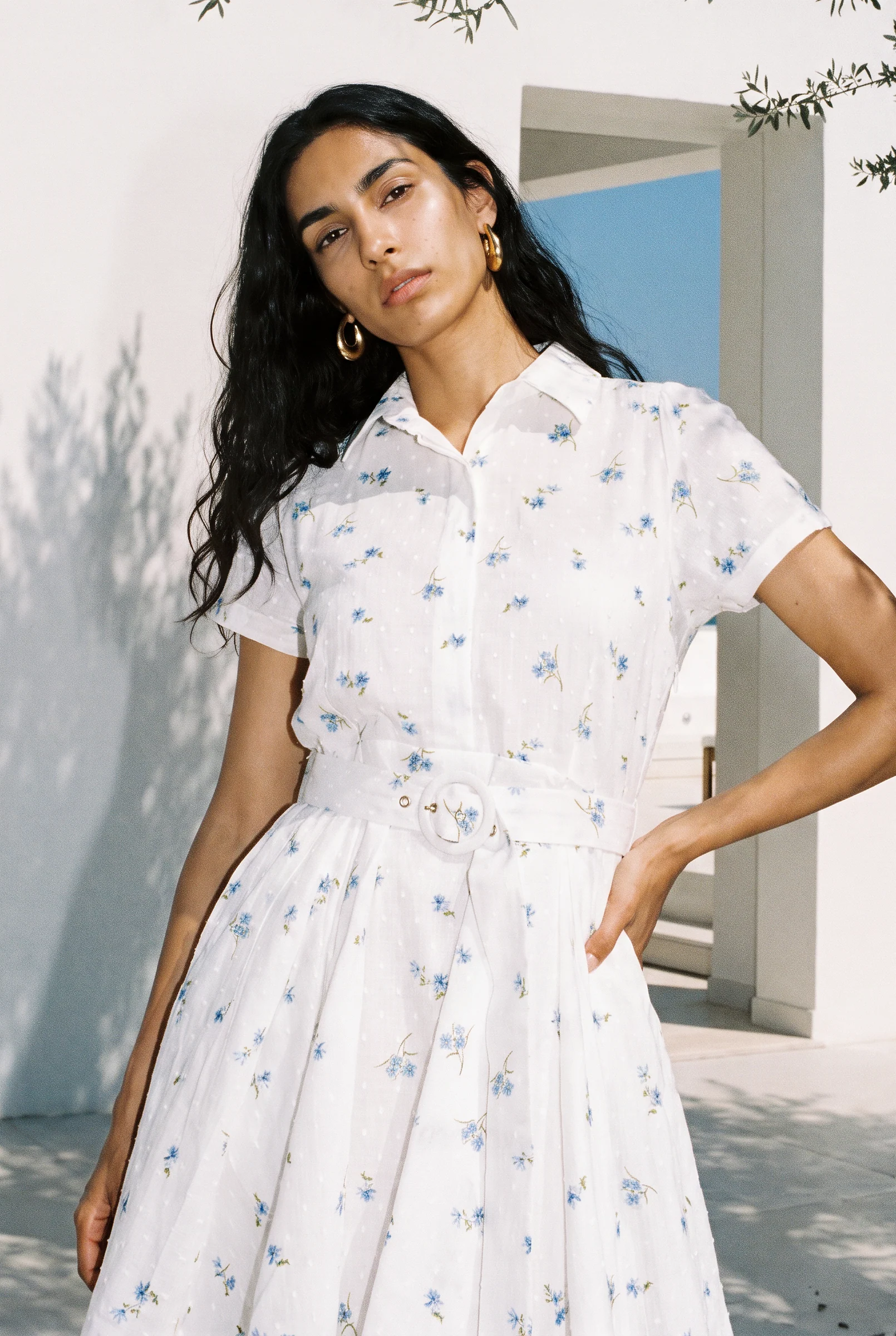 A model in a white floral shirt dress, hand on hip, against a white wall in sunlight
