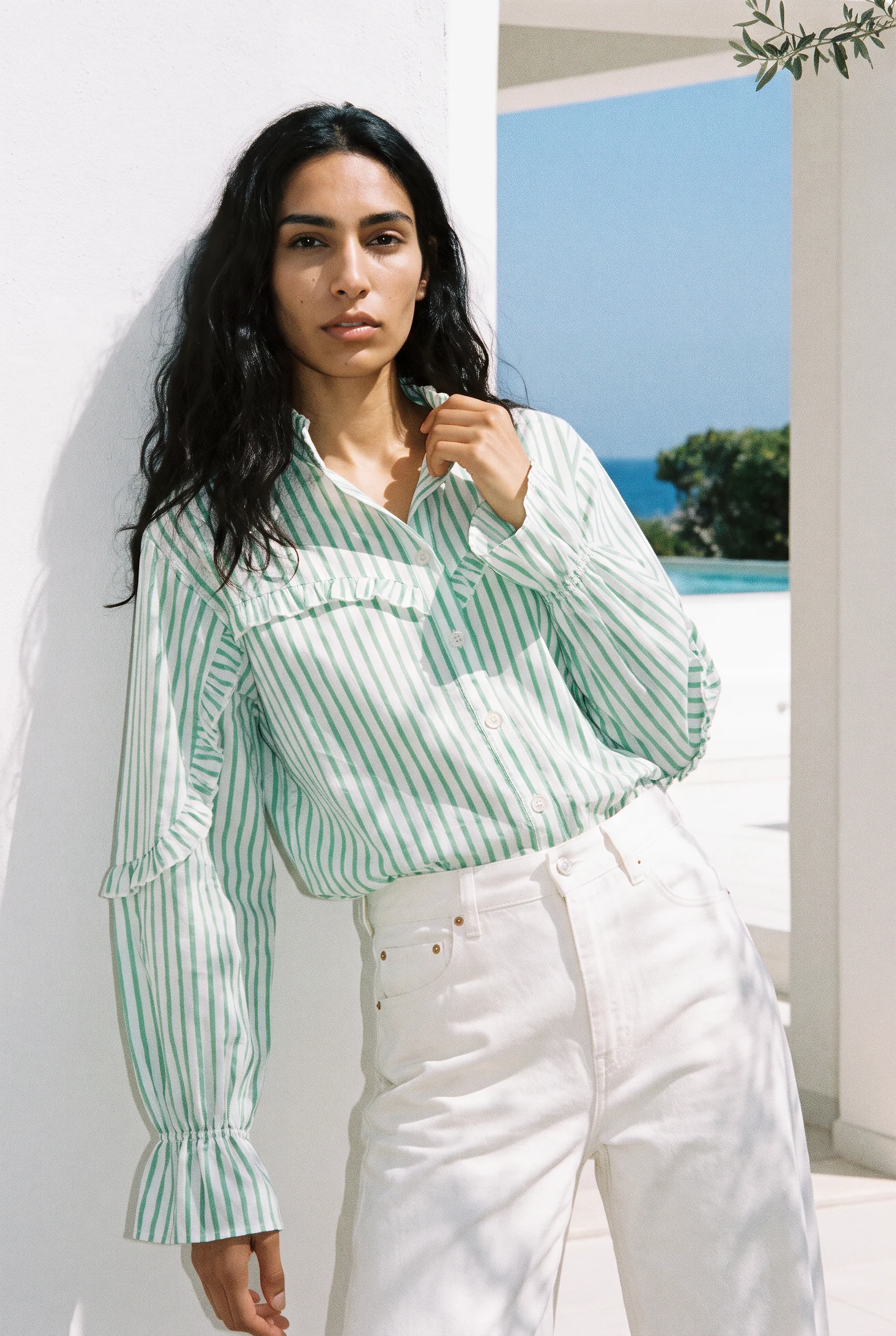A model in a mint striped shirt with a ruffled yoke and white jeans, against a white wall by the sea