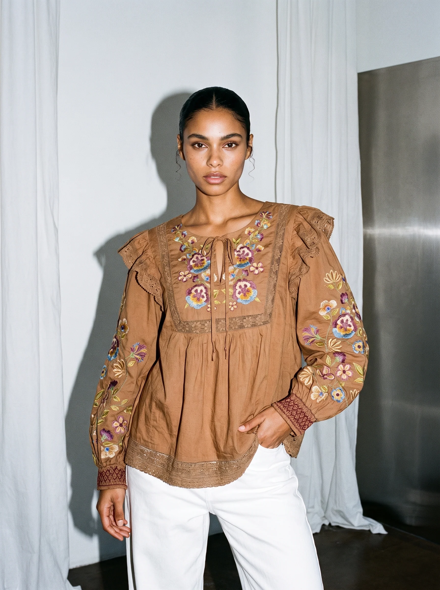 A model in the embroidered tan blouse and white trousers in a studio with a white curtain
