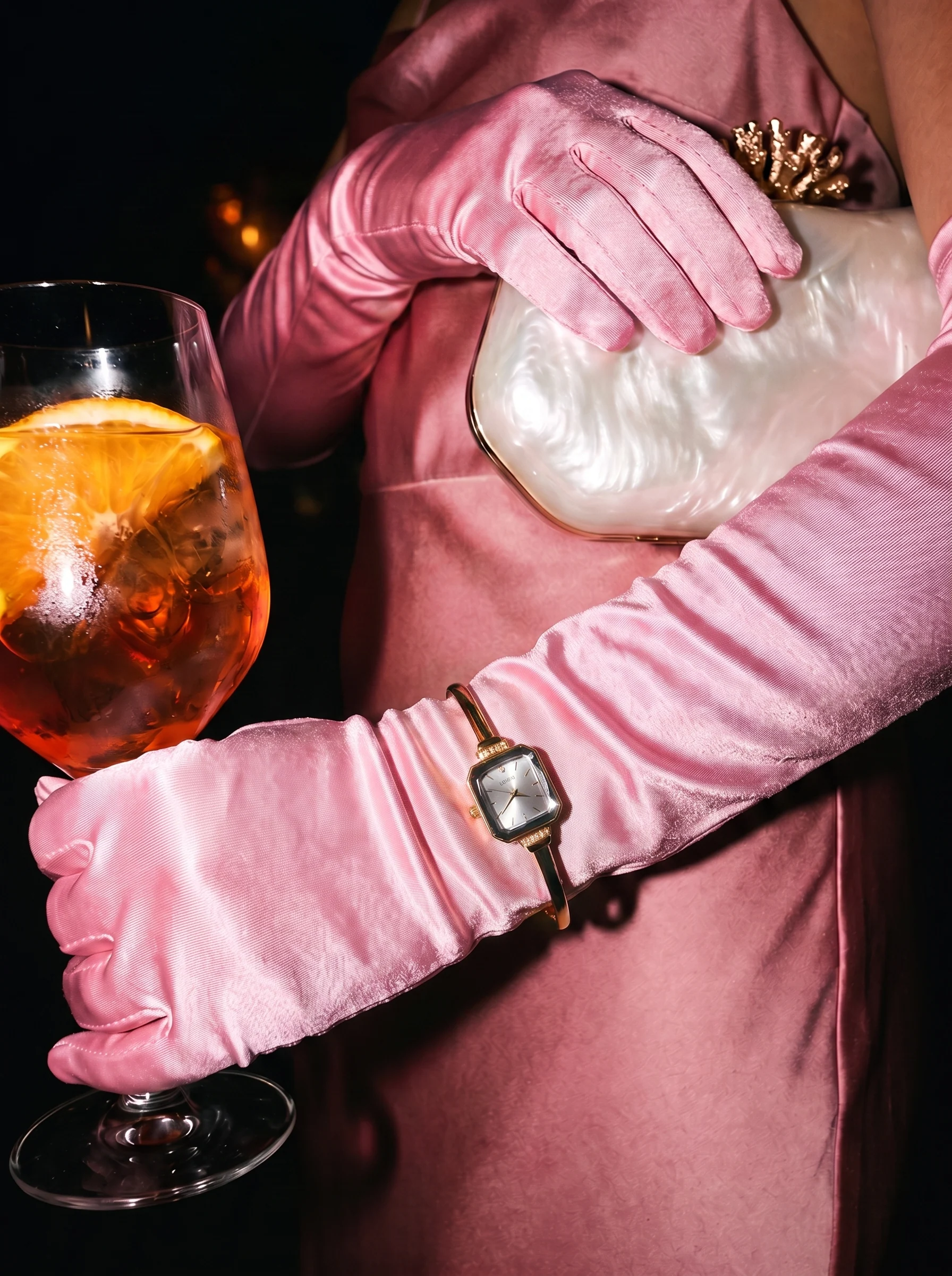 A woman in pink satin and long gloves, a spritz in one hand and a pearl clutch on her lap, the watch at her wrist