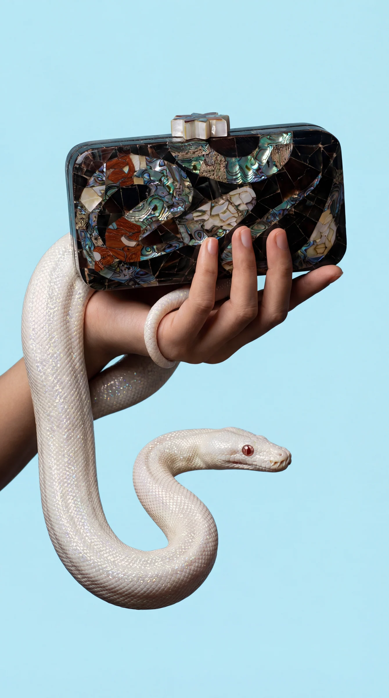 A hand holding a mother-of-pearl mosaic clutch, a white snake coiled around the wrist, on a pale blue ground